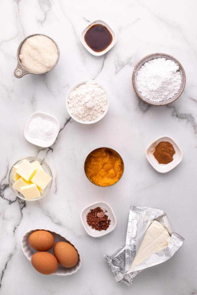 Overhead view of pumpkin roll ingredients including flour, sugar, eggs, butter, pumpkin puree, cream cheese, and spices arranged on a marble surface.