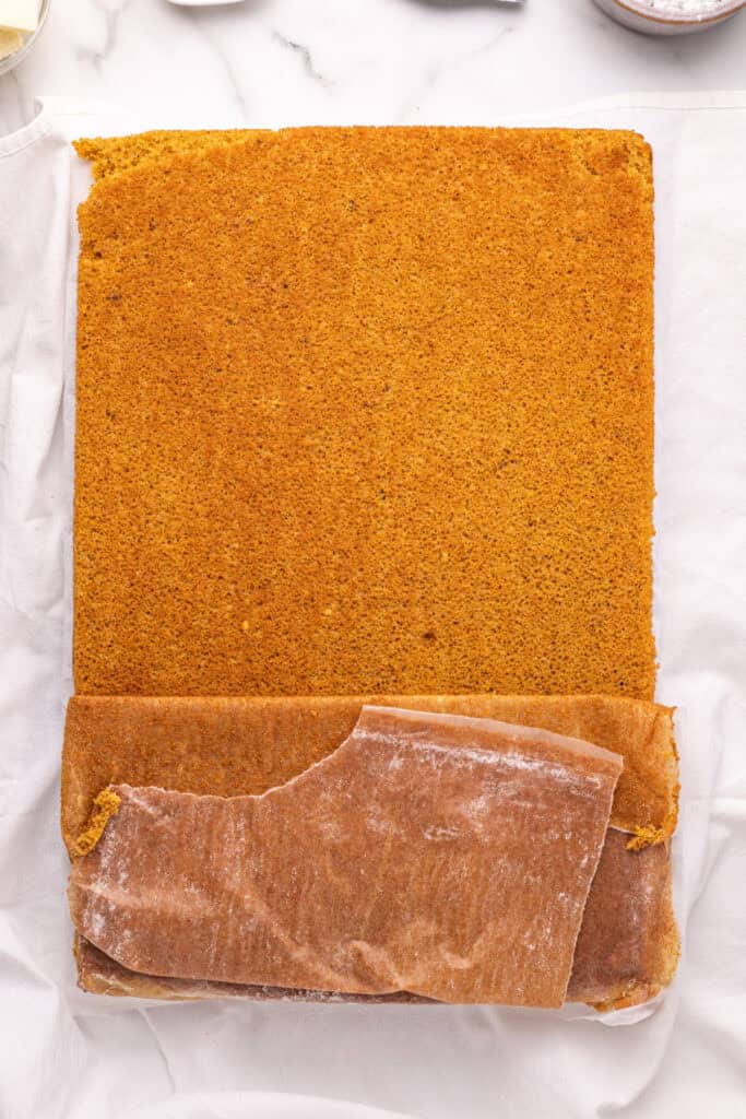 Pumpkin roll cake inverted onto a kitchen towel, with parchment paper being peeled away.