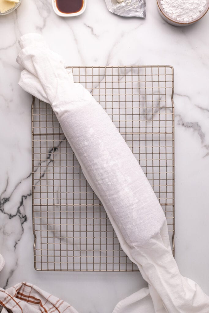 Pumpkin roll wrapped in a sugared kitchen towel cooling on a wire rack.