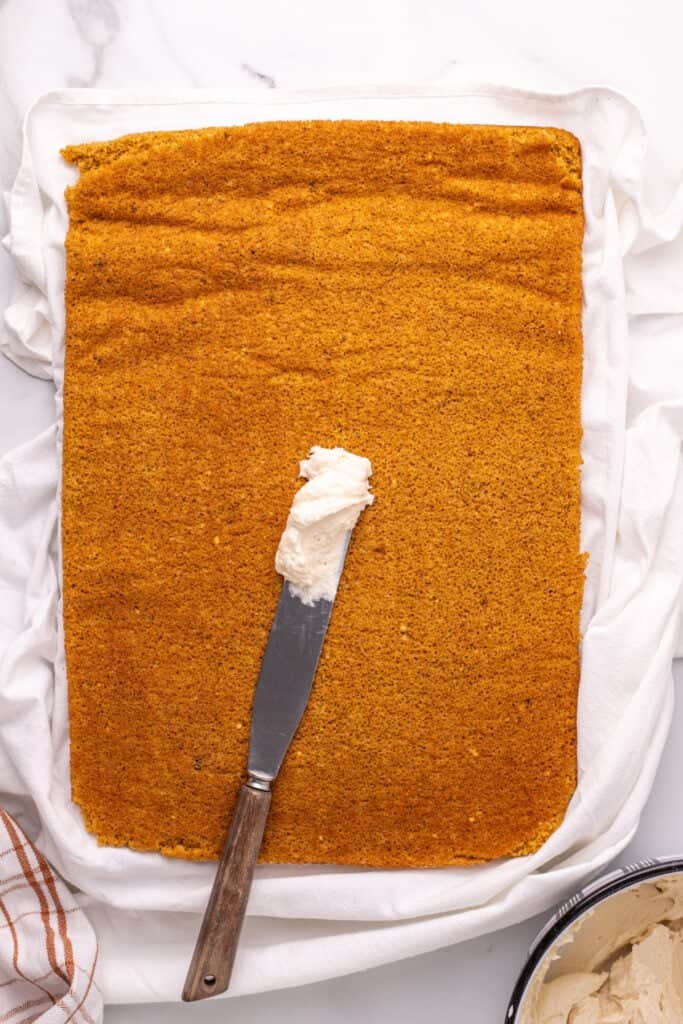 Pumpkin roll cake with offset spatula spreading cream cheese filling.