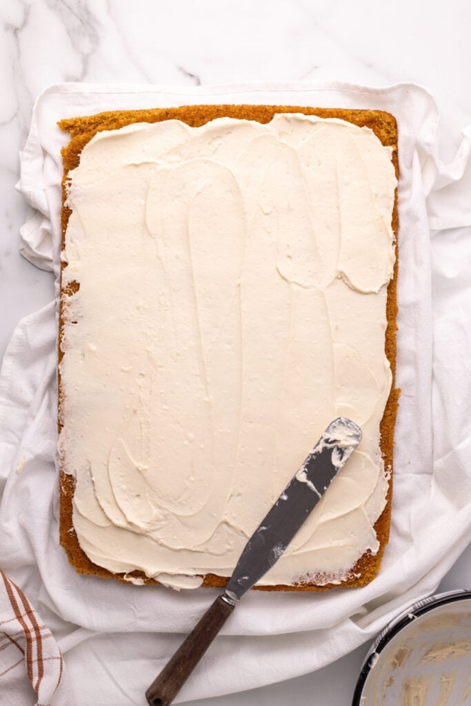 Pumpkin roll cake fully covered in cream cheese filling with spatula on top.