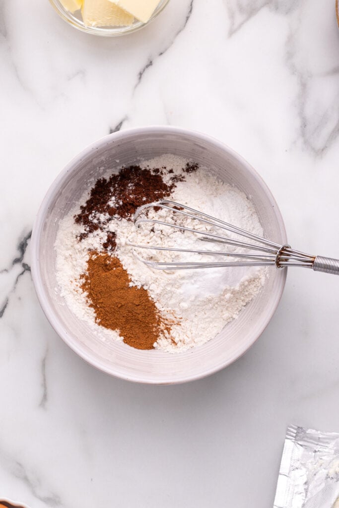 Mixing bowl with flour, cinnamon, cloves, baking powder, and baking soda being whisked together.