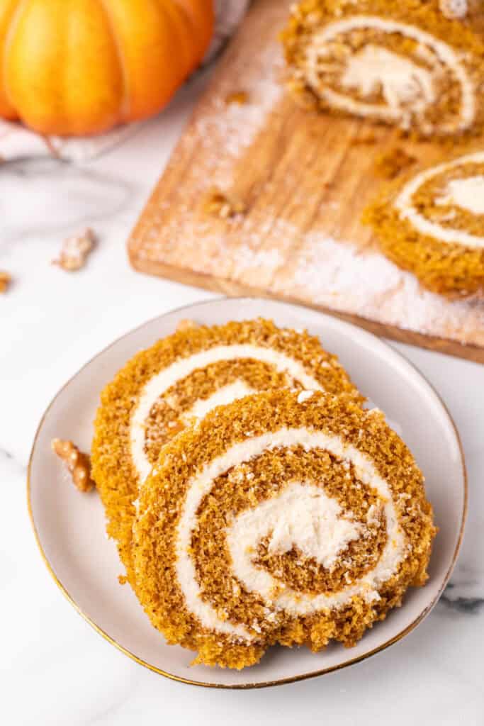 Pumpkin roll slices on a white plate with wooden board and pumpkin in background.
