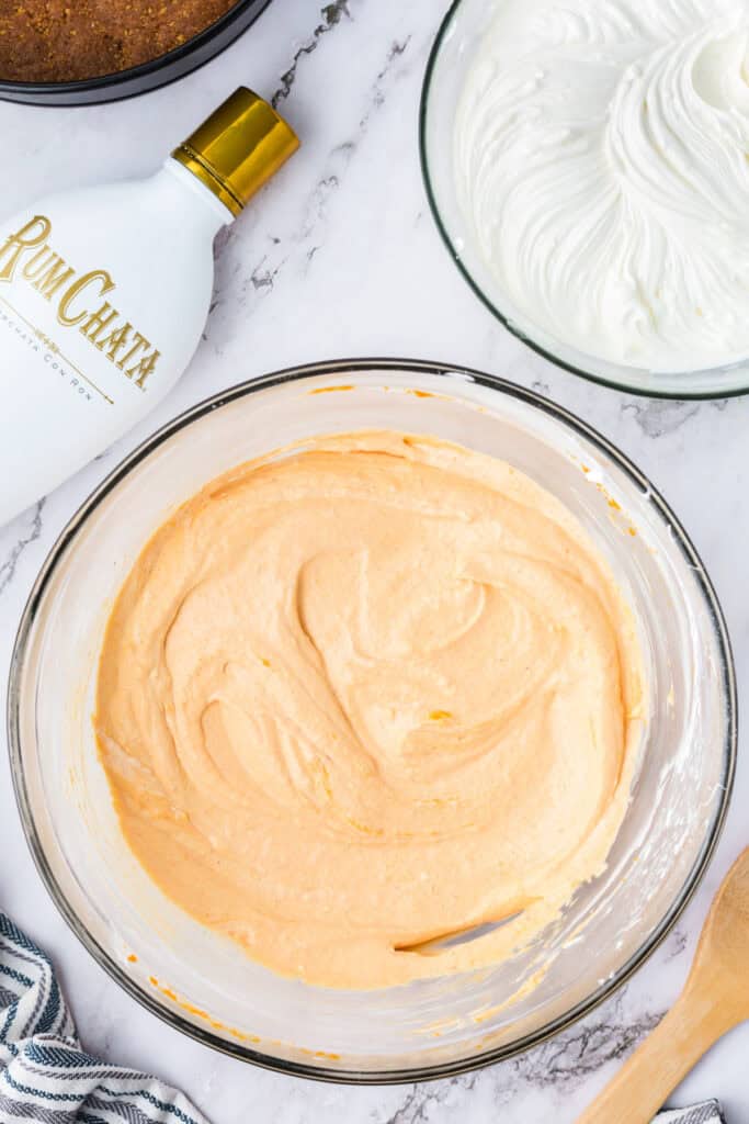 Bowl of pumpkin cheesecake filling mixed until smooth and creamy, with RumChata bottle, whipped mixture, and springform crust nearby.
