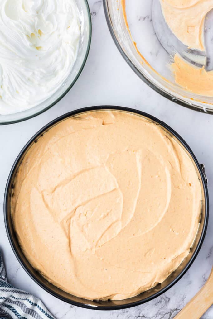 Pumpkin cheesecake filling layer spread evenly over the graham cracker crust in a springform pan, with whipped mixture in a bowl nearby.