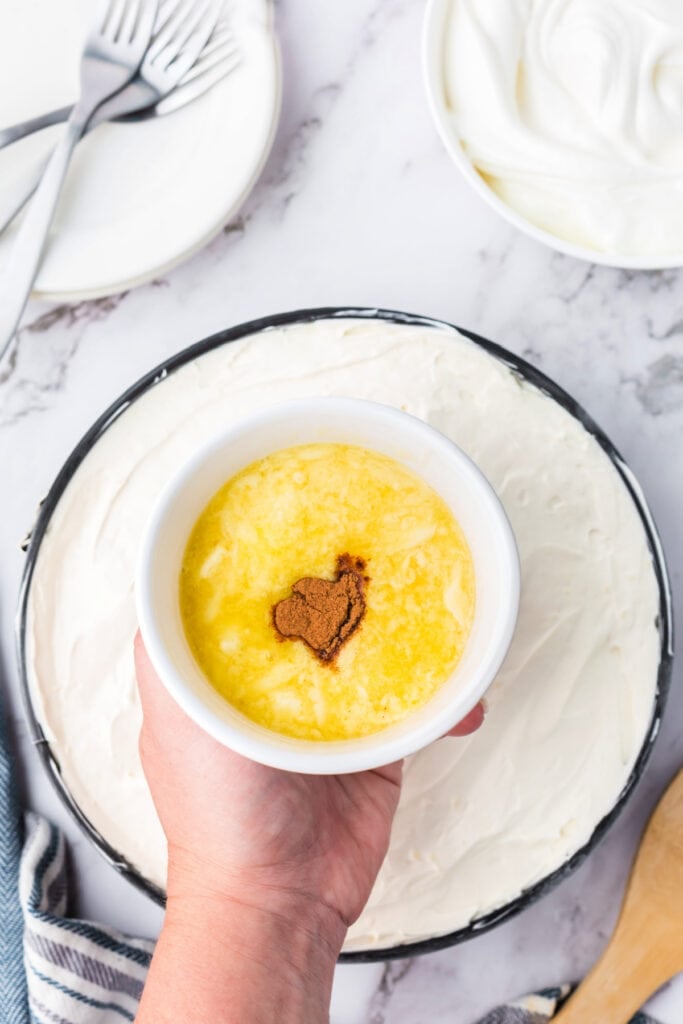 Hand holding a small bowl of melted butter and cinnamon mixture above the cheesecake, ready to make the cinnamon drizzle topping.
