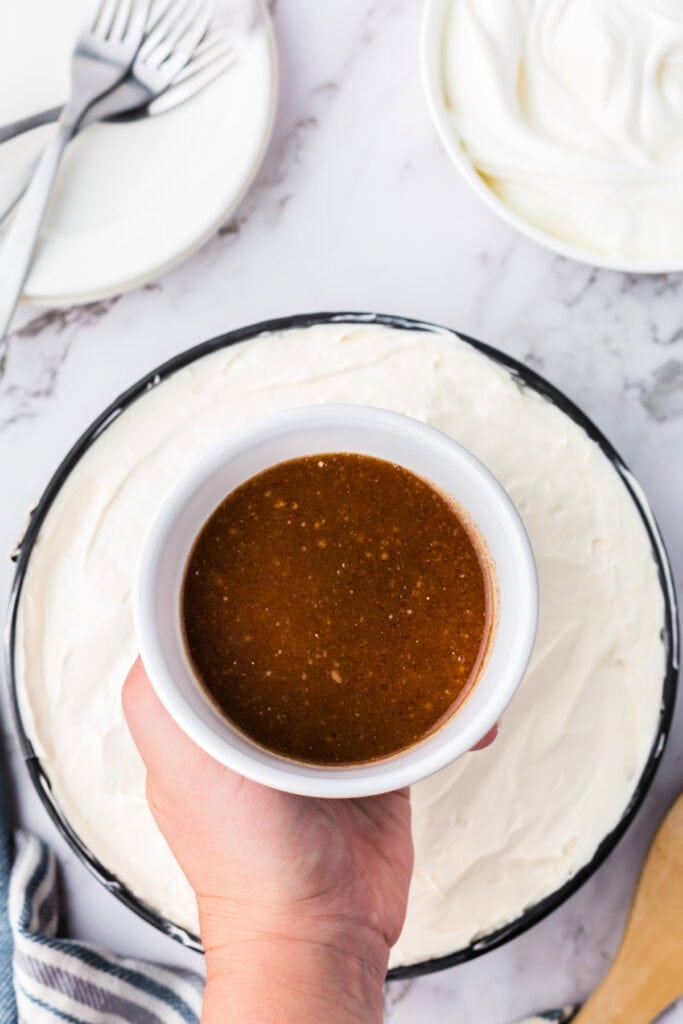 Hand holding a bowl of prepared cinnamon-brown sugar drizzle topping over the finished layered cheesecake.