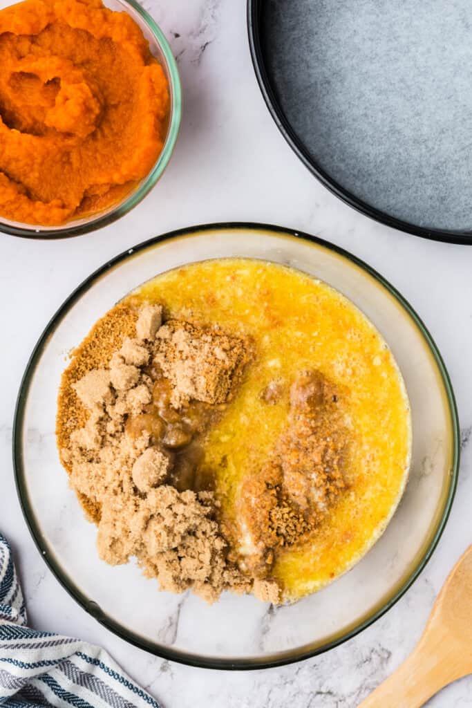 Mixing bowl filled with graham cracker crumbs, brown sugar, and melted butter for the cheesecake crust, with pumpkin puree and a lined springform pan nearby.