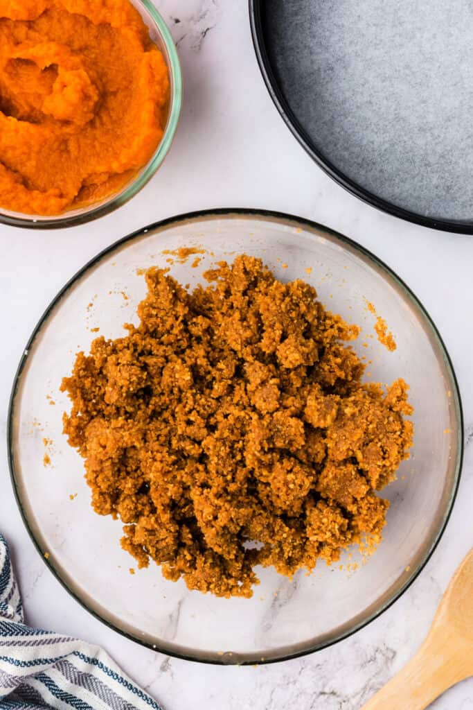 Prepared graham cracker crust mixture after being stirred together in a clear bowl, with pumpkin puree and springform pan in the background.