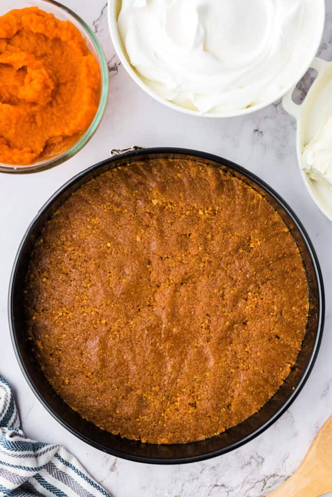 Smooth and evenly pressed graham cracker crust set in the springform pan, with pumpkin puree and cool whip bowls nearby.