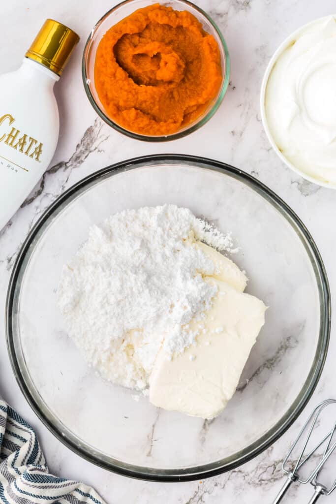 Mixing bowl with softened cream cheese and powdered sugar before blending, with pumpkin puree and RumChata bottle nearby.
