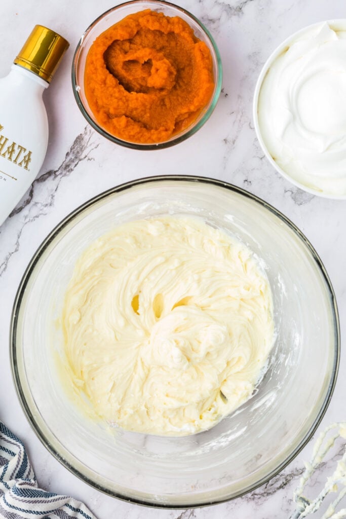 Cream cheese and powdered sugar mixture beaten smooth in a glass bowl, surrounded by pumpkin puree, RumChata, and cool whip.