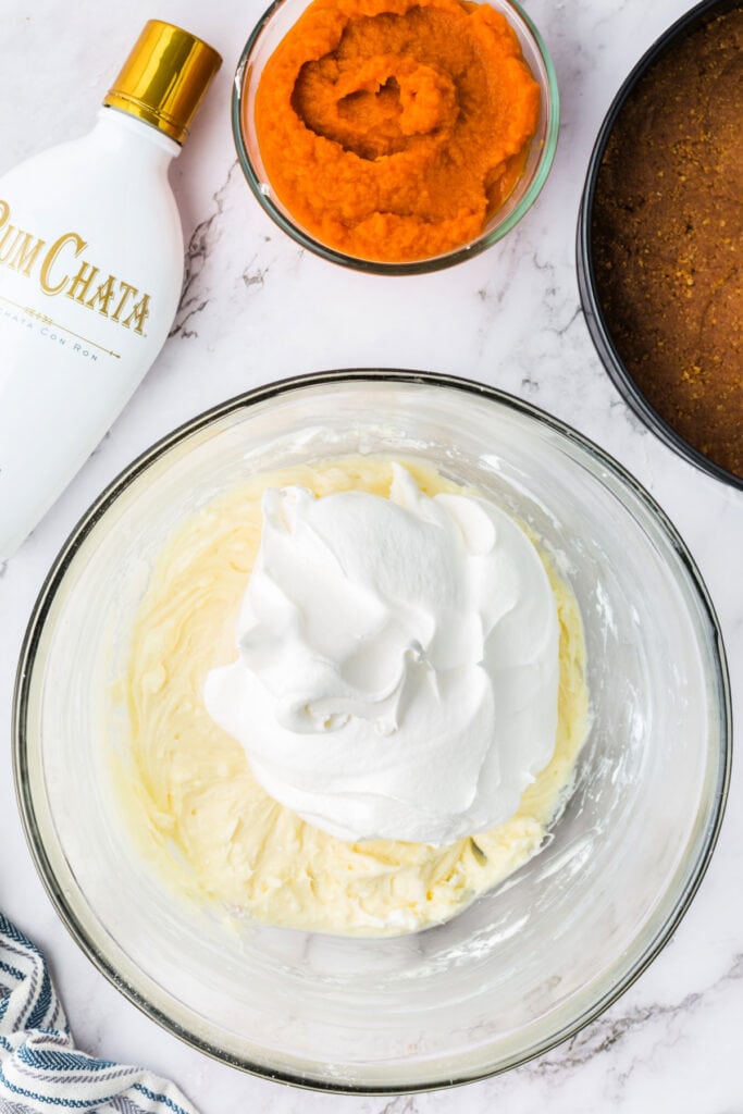 Bowl of cream cheese mixture topped with a large scoop of cool whip, ready to be folded together for the cheesecake filling.