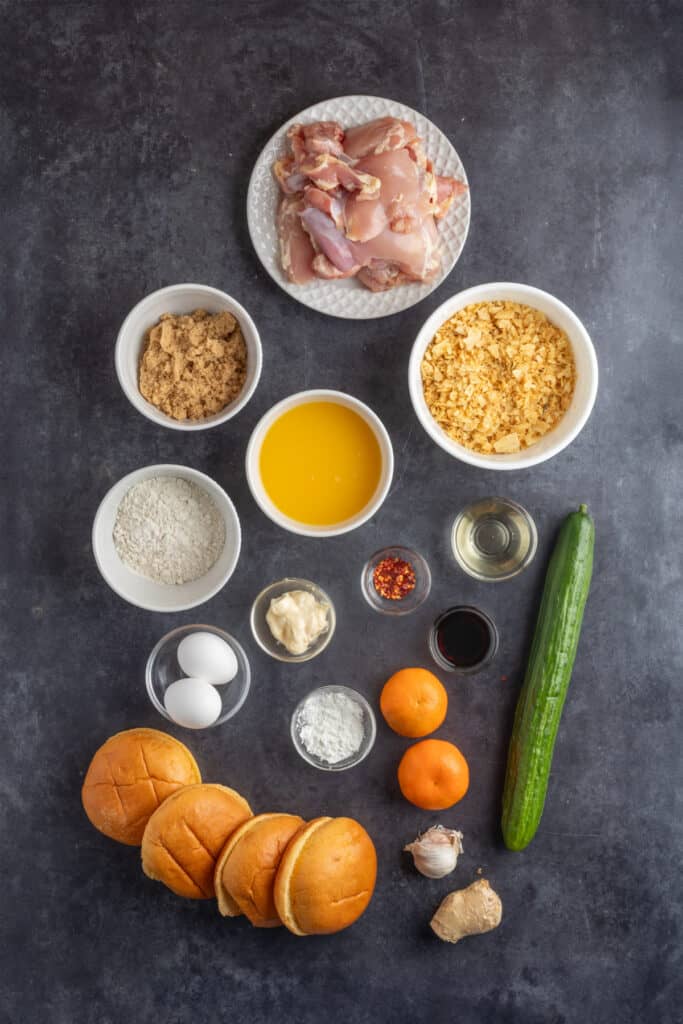 Overhead view of orange chicken sandwich ingredients including chicken thighs, flour, brown sugar, orange juice, cucumber, buns, spices, and condiments arranged on a dark surface.
