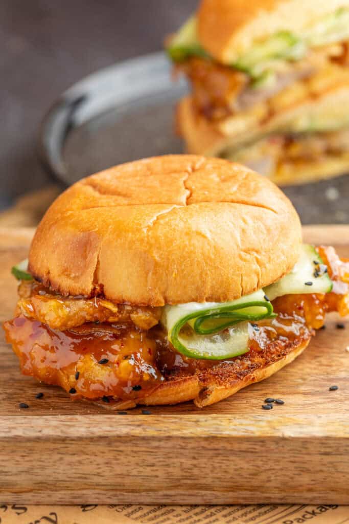 Close-up of a finished orange chicken sandwich with sauced chicken and cucumber ribbons on a toasted bun.