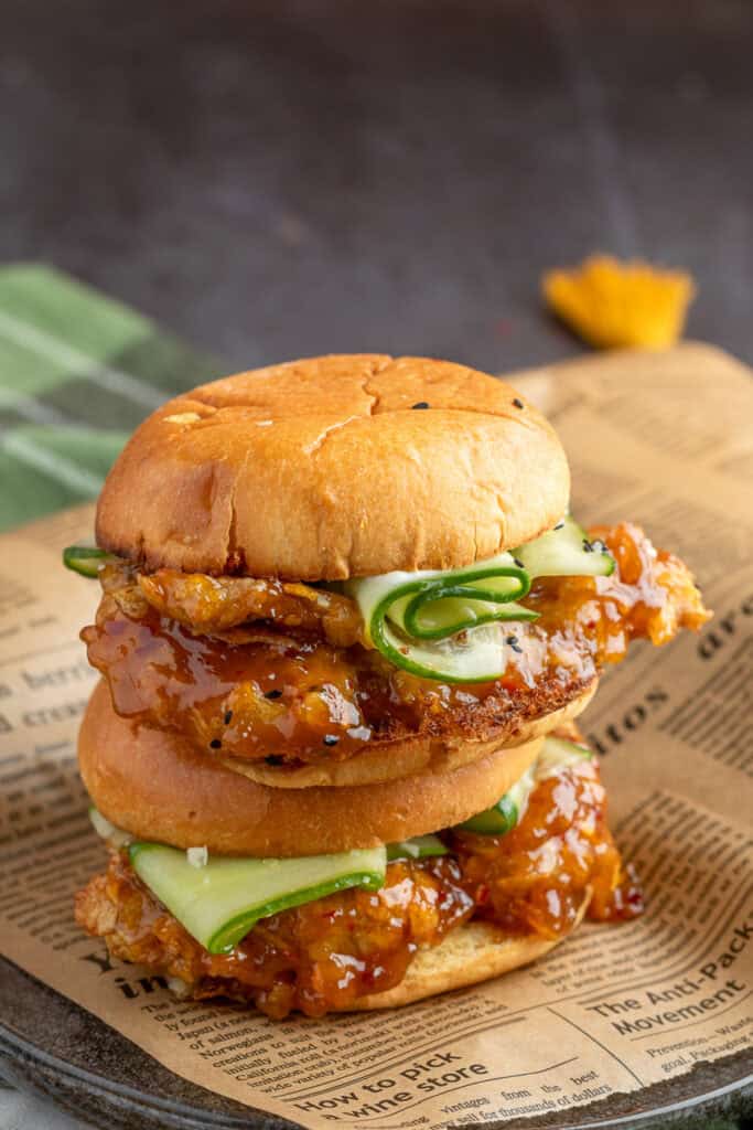 Stack of two orange chicken sandwiches layered with glazed chicken, cucumber ribbons, and sesame seeds on toasted buns.