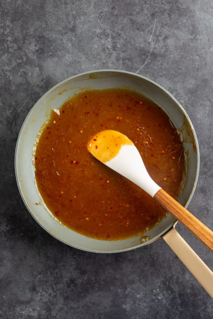 Saucepan filled with homemade orange sauce simmering with visible red pepper flakes, stirred with a white spatula.