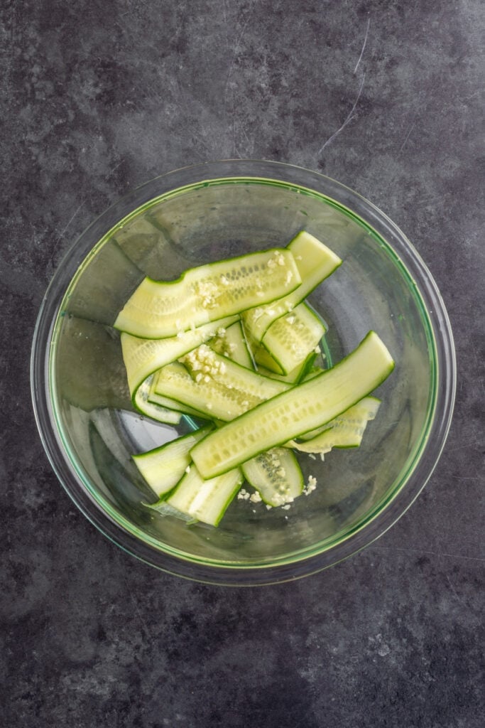 Glass bowl of cucumber ribbons tossed with minced garlic and rice vinegar for a quick cucumber salad.