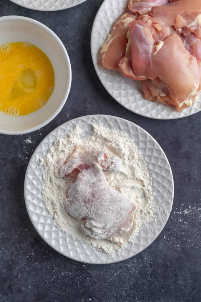 Raw chicken thigh coated in flour on a white plate next to beaten egg wash and uncooked chicken thighs.