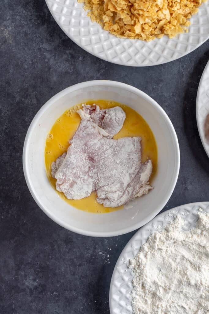 Chicken thigh dipped into beaten egg wash after dredging in flour, ready for coating.