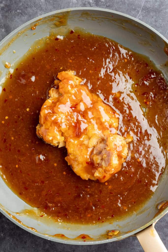 Breaded chicken thigh being coated in sticky homemade orange sauce inside a skillet.