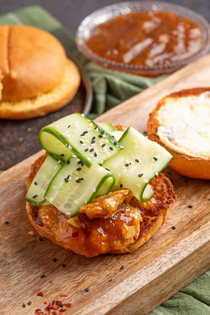 Open sandwich on a wooden board with sauced chicken, mayonnaise on the bun, cucumber ribbons, and sprinkled sesame seeds.