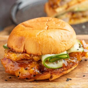 Square image of an orange chicken sandwich with saucy chicken, cucumber ribbons, and sesame seeds on a toasted bun.