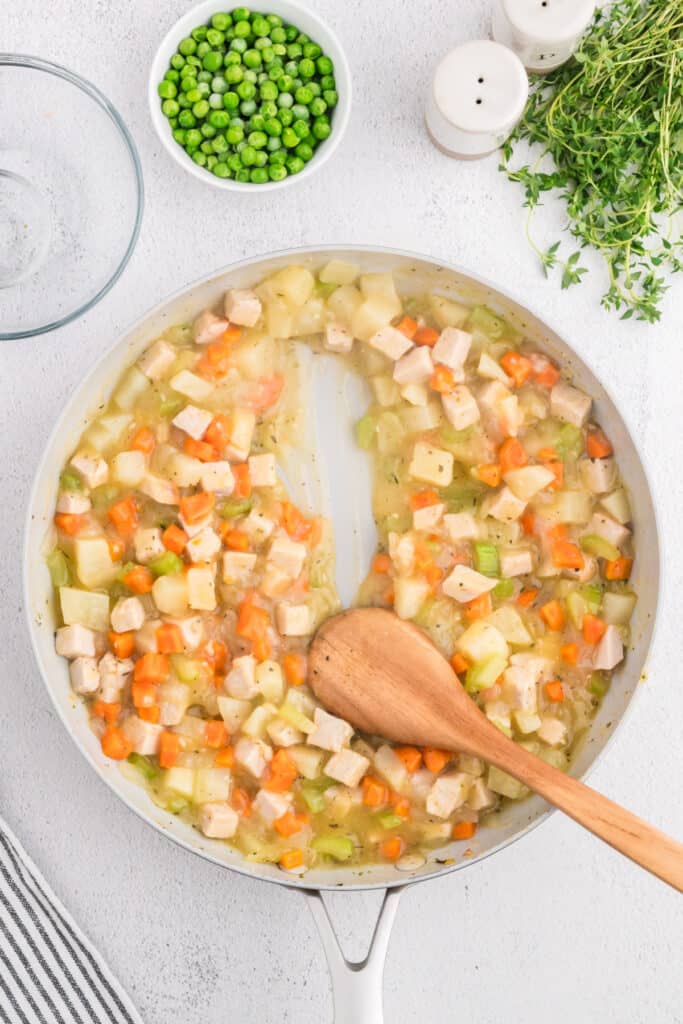 Skillet of creamy turkey pot pie filling being stirred with a wooden spoon, peas waiting to be added.