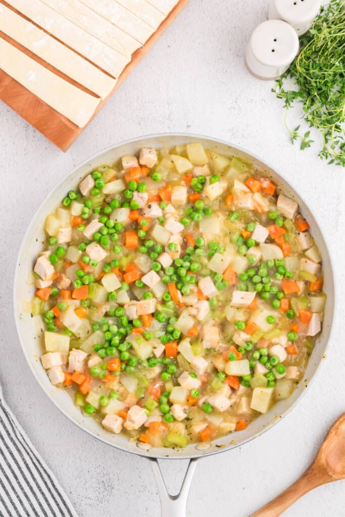 Turkey pot pie filling in skillet with peas mixed in, puff pastry strips ready on a cutting board.