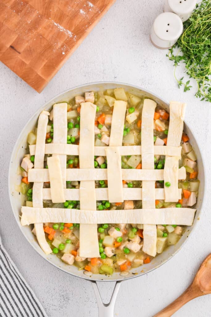 Skillet with puff pastry lattice partially completed over turkey pot pie filling.