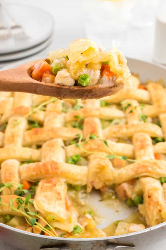 Wooden spoon lifting a serving of puff pastry turkey pot pie with turkey, peas, carrots, and flaky crust.