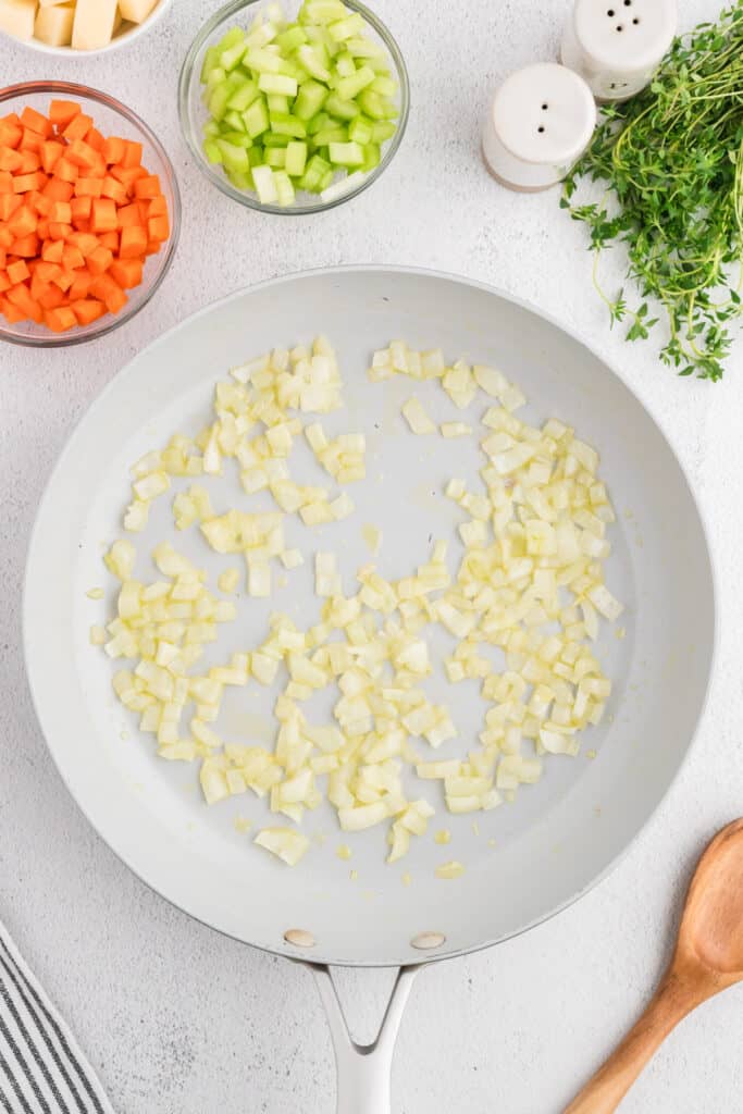 Onions sautéing in a white skillet with olive oil, surrounded by bowls of chopped vegetables.