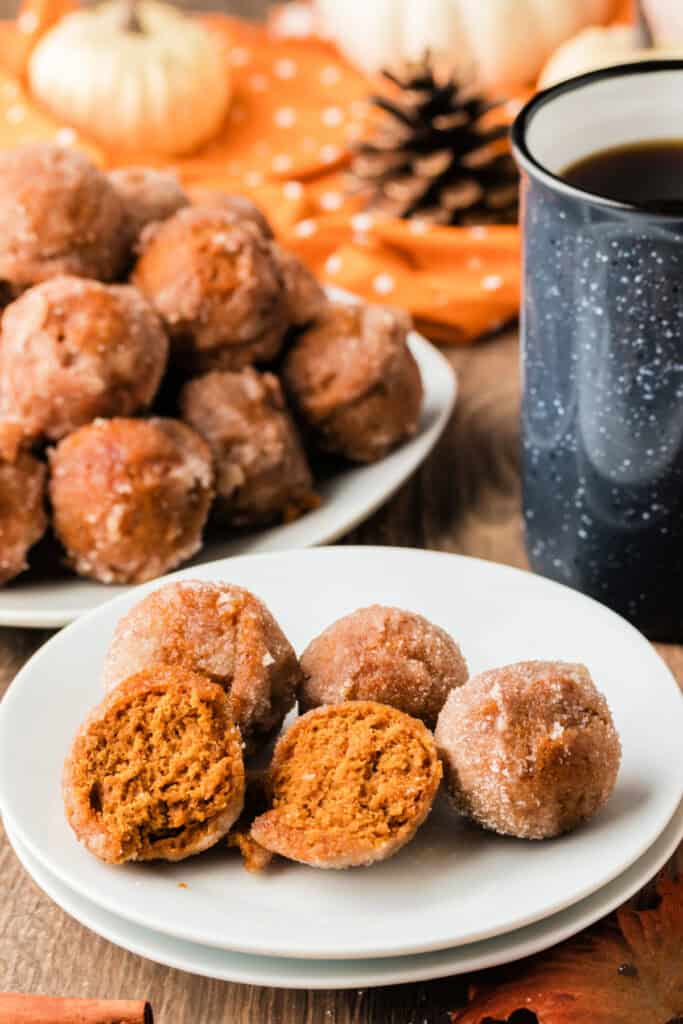 Plate of pumpkin donut holes coated in cinnamon sugar, with one cut open to show the soft pumpkin spice center, next to a coffee mug.