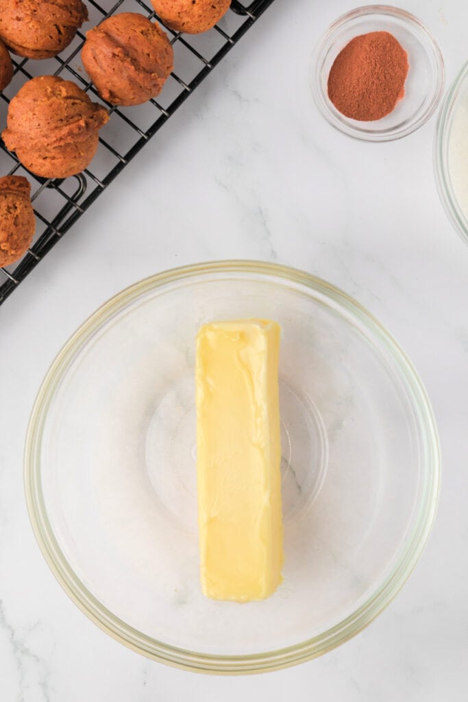 Stick of butter in a glass bowl next to baked pumpkin donut holes on a cooling rack.