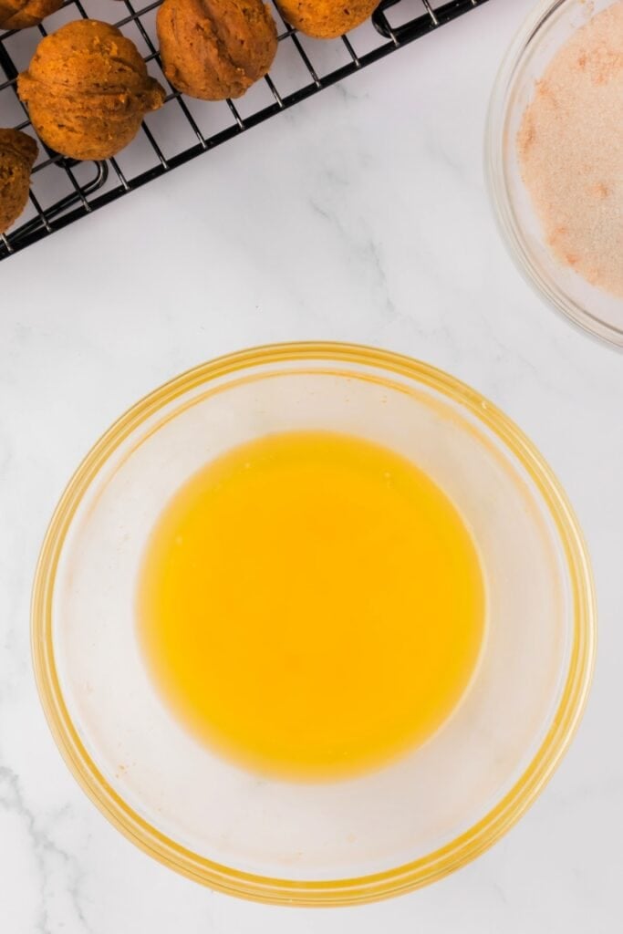 Melted butter in a glass bowl next to baked pumpkin donut holes and a bowl of cinnamon sugar.