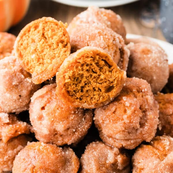 Pile of cinnamon sugar pumpkin donut holes stacked in a bowl, one cut in half on top to show the moist pumpkin dough inside.