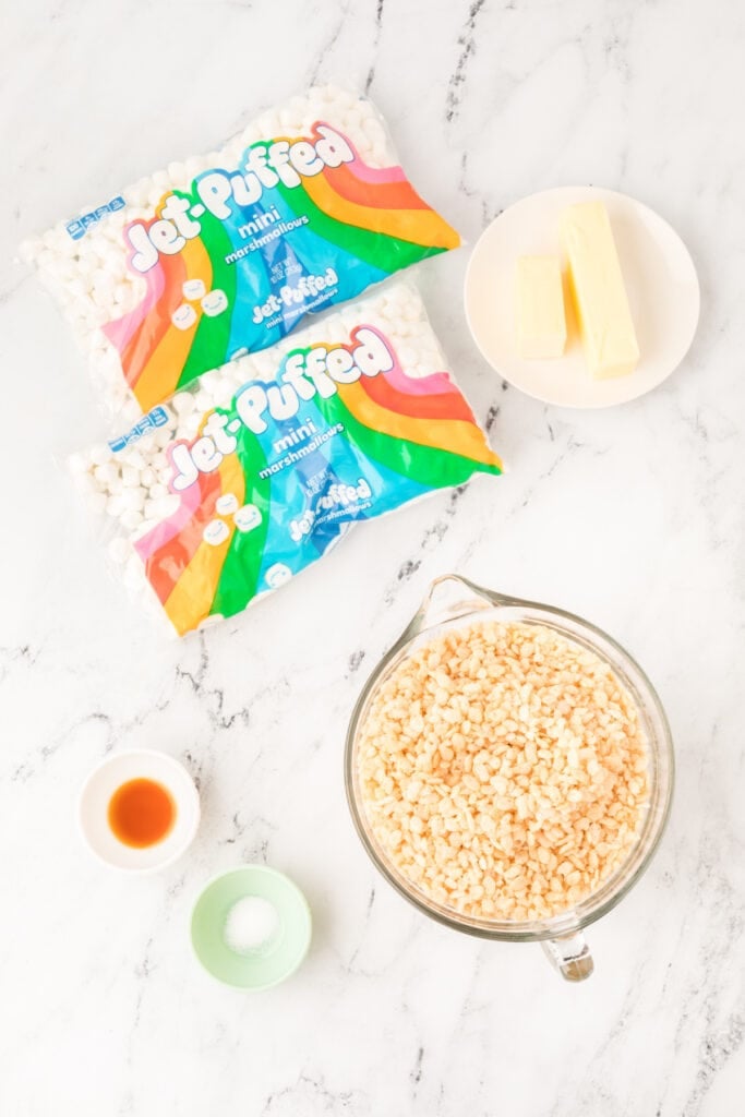 Ingredients for Browned Butter Rice Crispy Treats including mini marshmallows, butter sticks, crispy rice cereal, vanilla extract, and salt on a marble surface.