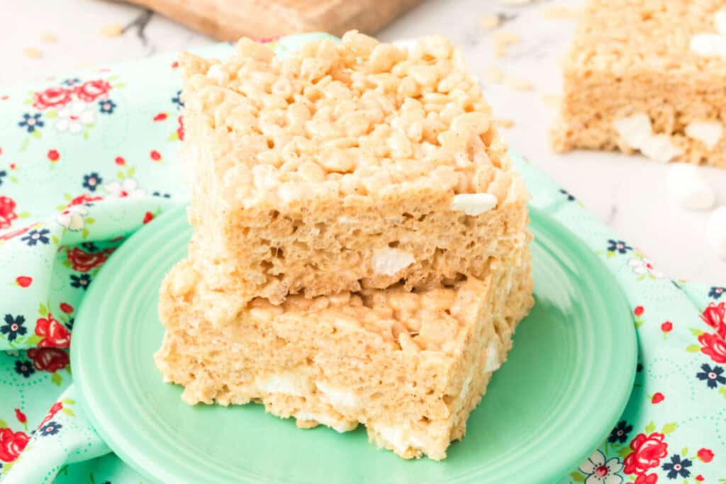 Close-up of two Browned Butter Rice Crispy Treats stacked on a green plate with a floral cloth and extra bars in the background.