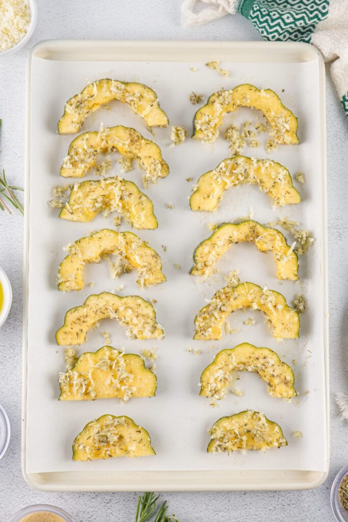Uncooked acorn squash slices on a parchment-lined baking sheet topped with parmesan and herbs.