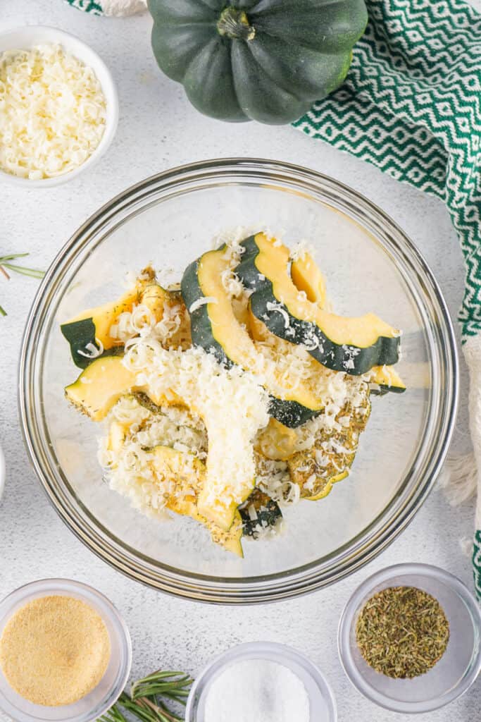 Bowl of acorn squash slices topped with shredded parmesan cheese and herbs before roasting.
