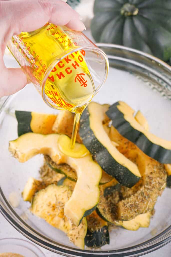 Olive oil being poured over acorn squash slices in a mixing bowl with herbs and spices.