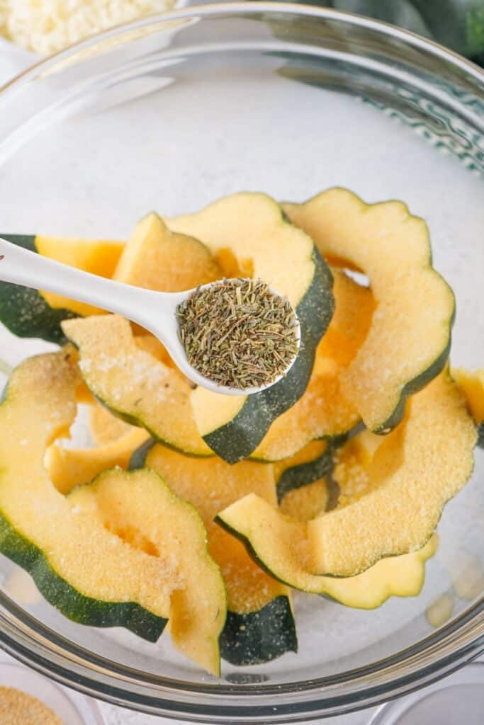 Spoonful of herbes de provence being added to sliced acorn squash in a mixing bowl.