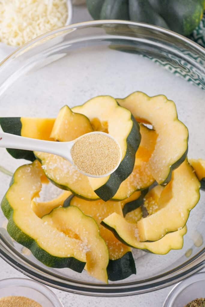 Spoonful of garlic powder being sprinkled over acorn squash slices in a mixing bowl.