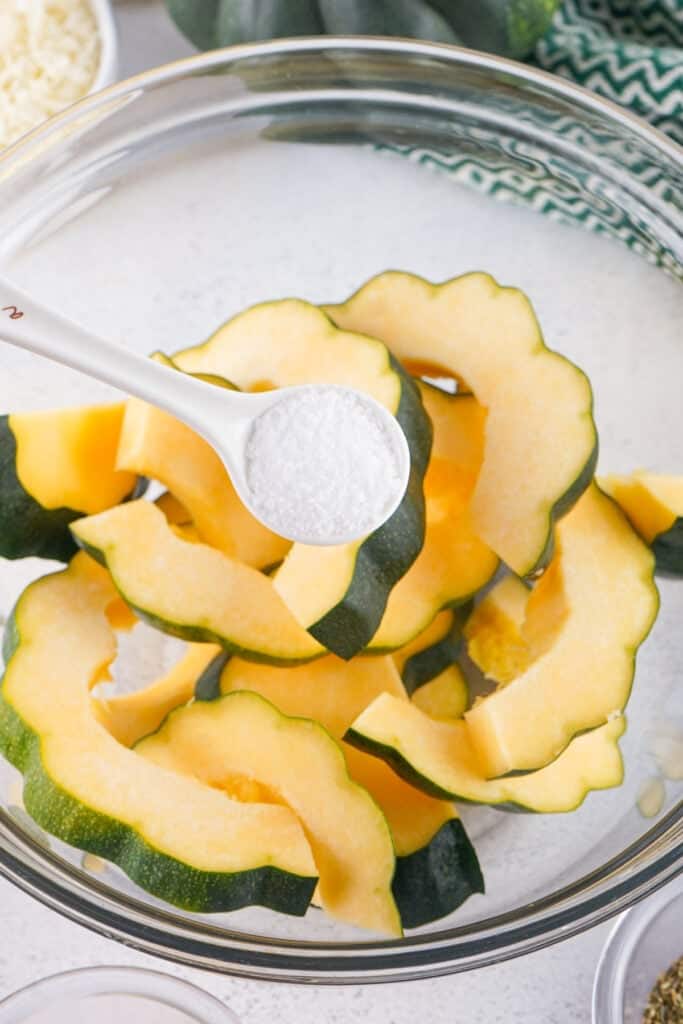 Spoonful of coarse salt being added to sliced raw acorn squash in a mixing bowl.