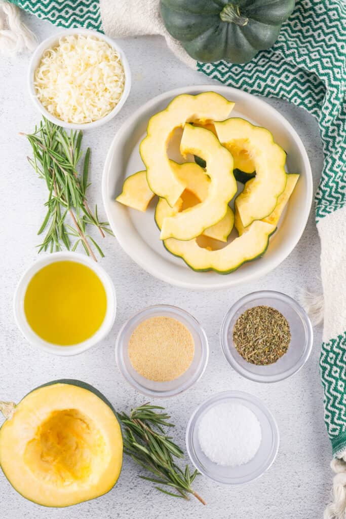 Overhead shot of acorn squash slices with olive oil, parmesan, garlic powder, herbs, and salt arranged on a countertop.
