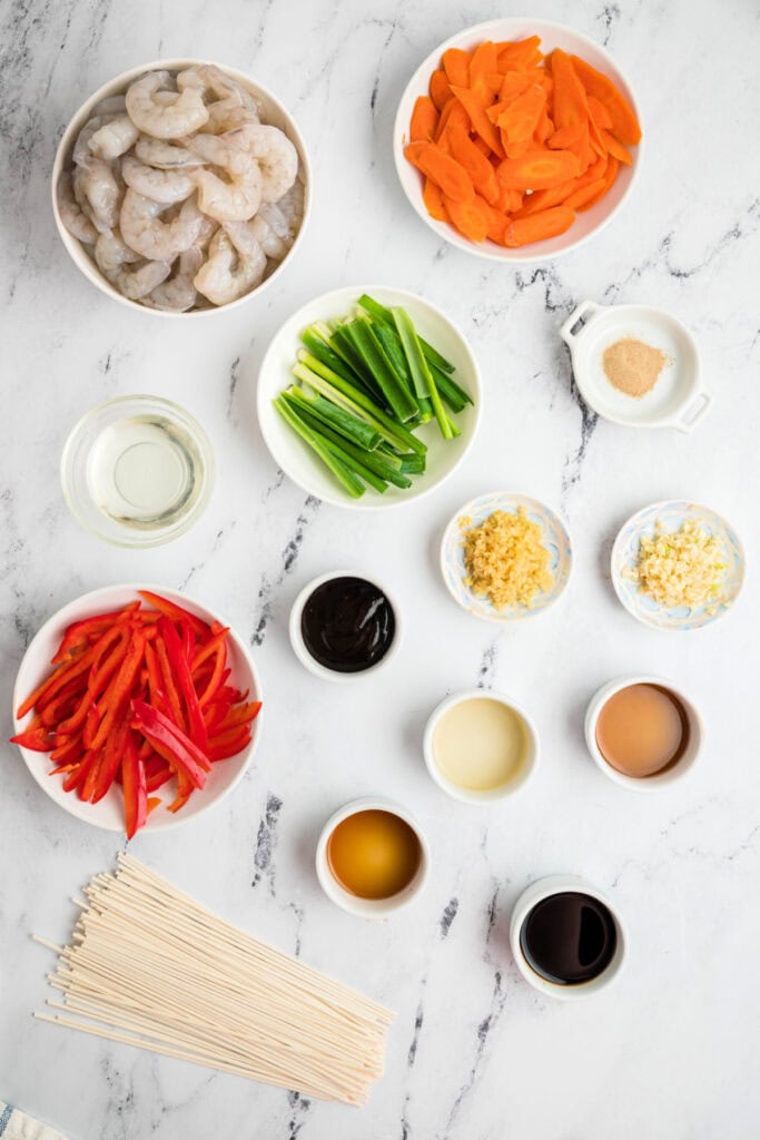 Overhead view of raw shrimp, sliced carrots, red bell pepper, green onions, garlic, ginger, and lo mein sauce ingredients laid out on a marble surface.