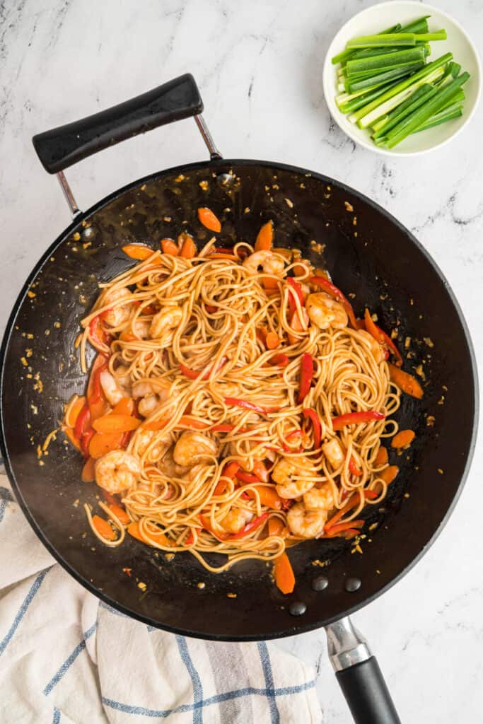 Shrimp, noodles, carrots, and bell peppers tossed together in a wok with sauce.