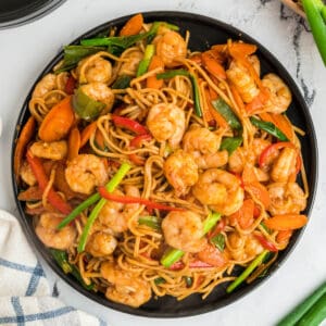 Square overhead image of shrimp lo mein with shrimp, noodles, and vegetables served on a black plate.