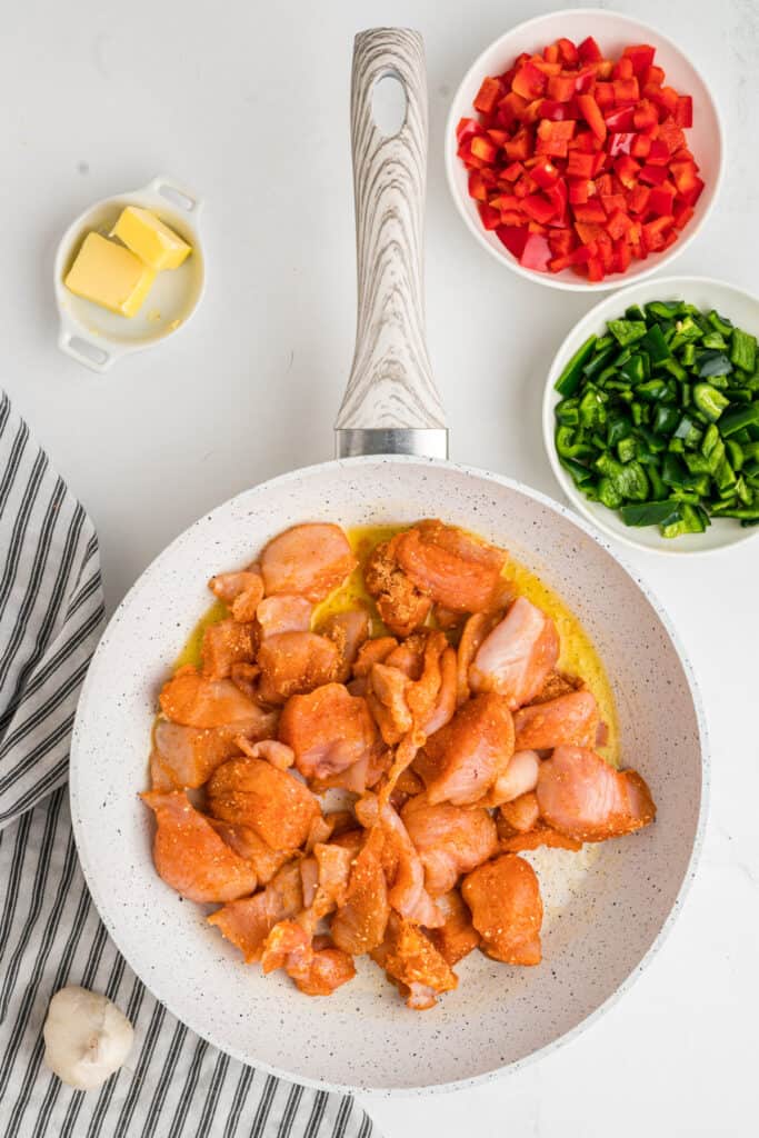 Seasoned chicken added to a skillet with bowls of butter and chopped peppers nearby.