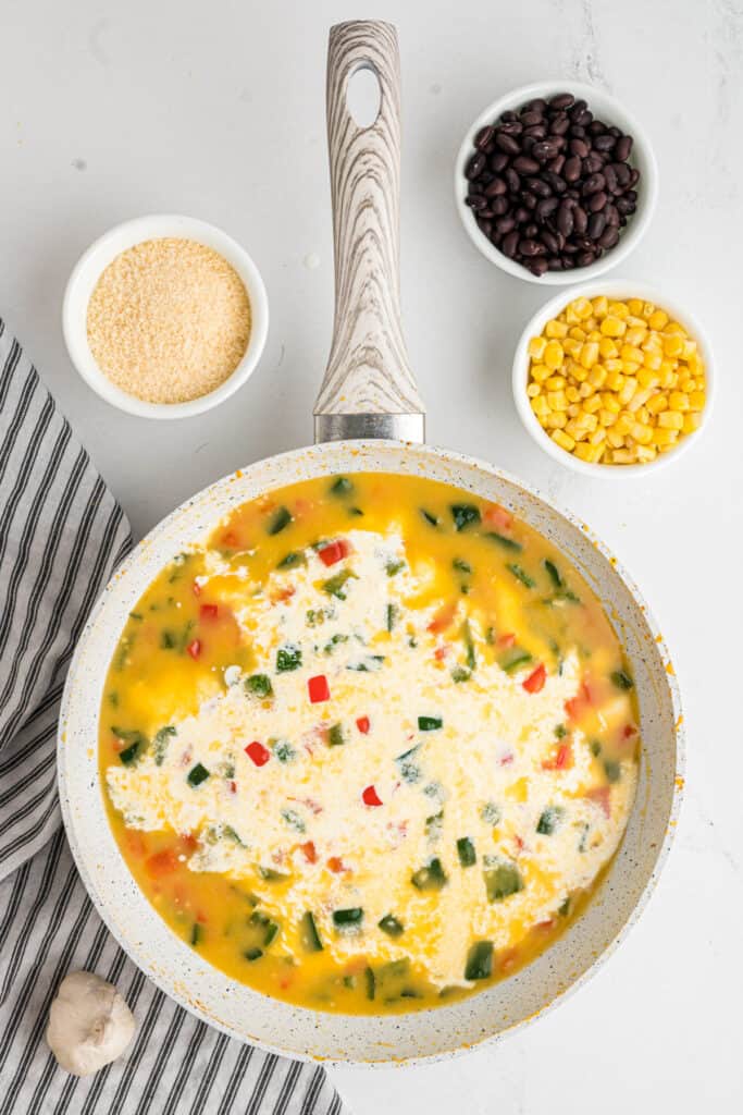 Cream poured into the skillet with peppers, cheese, beans, and corn nearby.